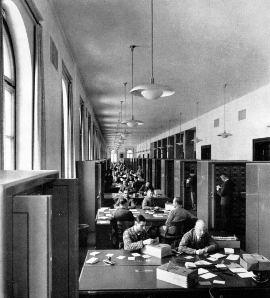 Großer Lesesaal mit langen Tischen, vielen Männern, hohen Fenstern und hängenden Lampen in einem historischen Gebäude