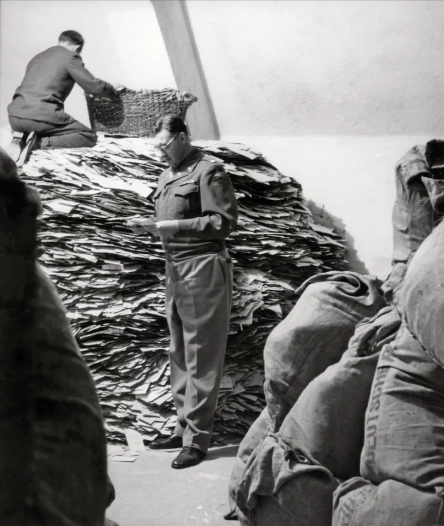 Zwei Männer in Uniform vor einem großen Stapel Papier und mehreren Säcken in einem Raum