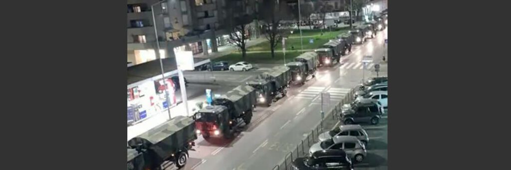 Das Foto zeigt einen langen Konvoi an Militärfahrzeugen, der über die Straße in einem Stadtgebiet fährt. Die Fahrzeuge sind alle mit Tarnmustern bemalt und um sie herum stehen einige parkende Autos.