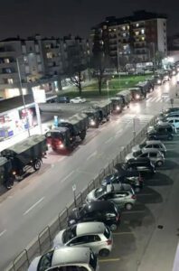 Das Foto zeigt einen langen Konvoi an Militärfahrzeugen, der über die Straße in einem Stadtgebiet fährt. Die Fahrzeuge sind alle mit Tarnmustern bemalt und um sie herum stehen einige parkende Autos.