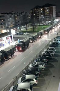 Das Foto zeigt einen langen Konvoi an Militärfahrzeugen, der über die Straße in einem Stadtgebiet fährt. Die Fahrzeuge sind alle mit Tarnmustern bemalt und um sie herum stehen einige parkende Autos.