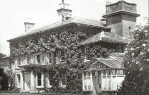 Eine Schwarzweißfotografie eines großen Hauses mit vielen Fenstern und einem kleinen Turm. Die gesamte Fassade ist mit dichten Kletterpflanzen bewachsen.