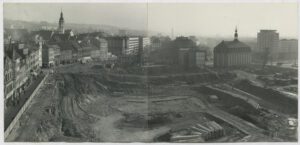 Eine Schwarzweißfotografie einer Stadtansicht. Im Zentrum des Bildes klafft allerdings eine große unbebaute Fläche, auf der wohl Bauarbeiten stattfinden.