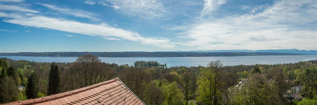 Eine Panoramaaufnahme von einem Dach aus. Zu sehen ist die Spitze des Daches, ein breiter grüner Waldstreifen und dahinter ein breites Gewässer. Hinter diesem folgen wieder Wald und schließlich die Berge. Der Himmel ist blau und vereinzelt mit Wolken bedeckt.