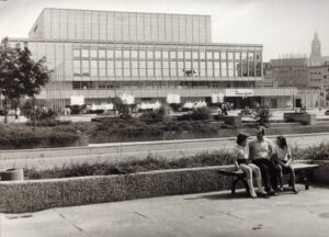 Eine Schwarzweißfotografie eines großen, kastenartigen Gebäudes mit einer großen Fensterfront. Im Vordergrund befinden sich einige Wasserspiel und Menschen auf einem Platz. In der unteren rechten Ecke sitzt ein Mann mit seinen zwei Kindern auf einer Straßenbank. Im Hintergrund sind eine Häuserfassade und ein hoher Kirchturm zu erkennen.