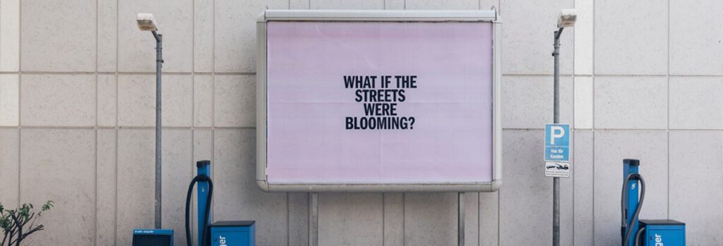 Vor einer grauen Wand hängt zwischen zwei Laternen eine große Werbetafel. Sie zeigt ein großes fliederfarbenes Plakat. Darauf ist in schwarzen Großbuchstaben zu lesen: "WHAT IF THE STREETS WERE BLOOMING?".
