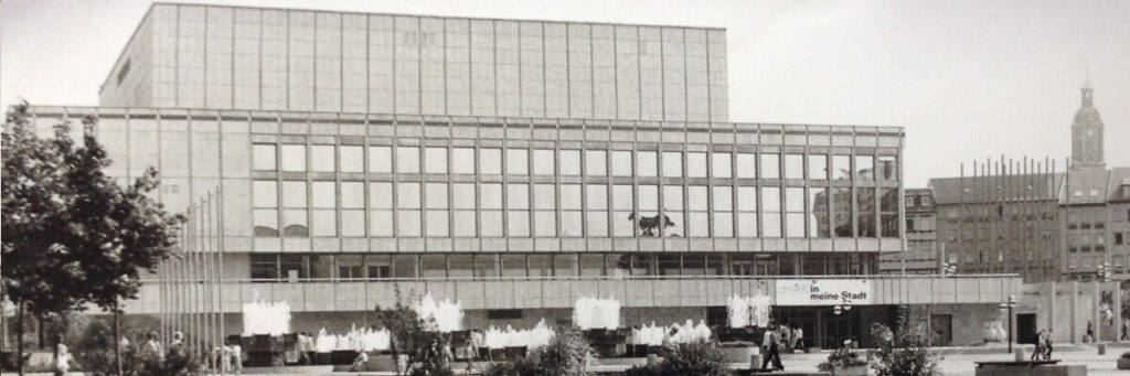 Eine Schwarzweißfotografie eines großen, kastenartigen Gebäudes mit einer großen Fensterfront. Auf dem Platz davor befinden sich einige Wasserspiel und einige Menschen. Im Hintergrund sind eine Häuserfassade und ein hoher Kirchturm zu erkennen.