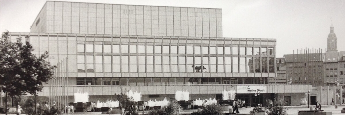 Eine Schwarzweißfotografie eines großen, kastenartigen Gebäudes mit einer großen Fensterfront. Auf dem Platz davor befinden sich einige Wasserspiel und einige Menschen. Im Hintergrund sind eine Häuserfassade und ein hoher Kirchturm zu erkennen.