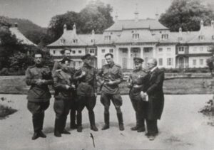 Die Schwarzweißfotografie zeigt eine Gruppe uniformierter Männer auf der Straße vor einem prächtigen Anwesen stehen.