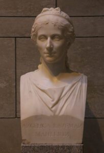 Die Büste einer Frau aus weißem Marmor. Sie ist jung und hat lange lockige Haare. Die Inschrift auf der Büste identifiziert sie als Angelica Kaufman. Dort ist außerdem das Wort "Mahlerin" eingraviert.