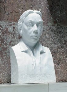 Die weiße, steinerne Büste einer Frau vor einer rötlich-grauen Steinwand. Die Inschrift auf der Büste identifiziert die Person als Käthe Kollwitz.