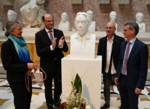 Eine Frau und drei Männer in Anzügen stehen um eine weiße Büste einer Frau herum. Vor der Büste wurden Blumen aufgestellt und einer der Männer applaudiert. Im Hintergrund sind weitere Büsten zu sehen.