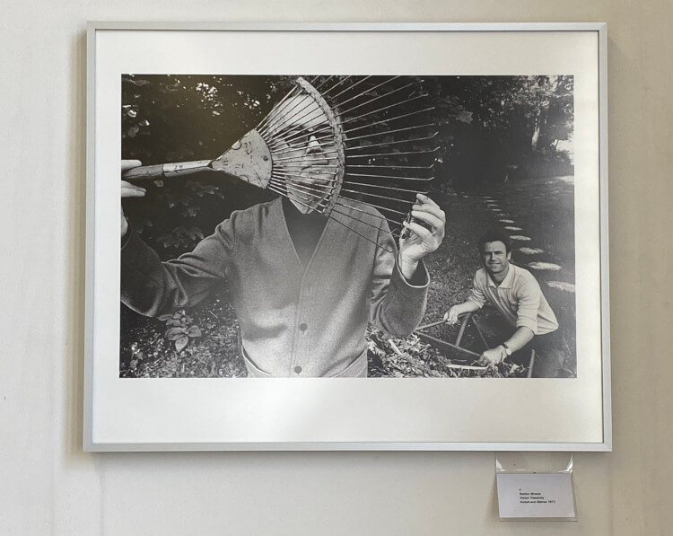 Eine Schwarzweißfotografie in einem silbernen Rahmen. Zu sehen sind zwei Männer in einem Garten. Einer kniet auf dem Boden während der andere steht und sich eine Laubrechen vors Gesicht hält. Unterhalb des Fotos wurde ein kleiner weißer Zettel mit Aufschrift angebracht.