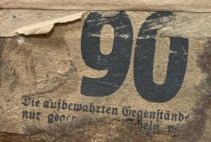 Ein abgenutztes Stück Papier mit einer großen aufgedruckten 90 und einem kurzen Text, der durch die Abnutzungen allerdings nicht mehr lesbar ist. Zu entziffern ist nur noch "Die aufbewahrten Gegenstände [...]".