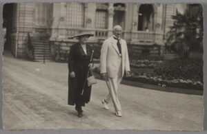 Auf der Schwarzweißfotografie laufen eine Frau und ein Mann über einen breiten Weg vor einem imposanten Gebäude und einem gut gepflegten Garten. Der Mann trägt einen weißen Anzug und eine dunkle Krawatte während die Frau ein schwarzes Kleid und einen großen Hut trägt.
