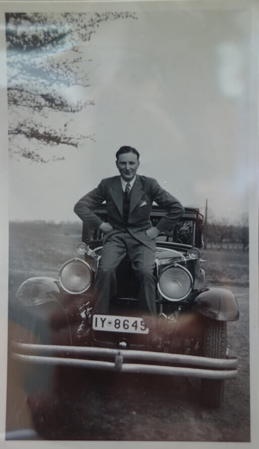 Schwarzweißfotografie eines jungen Mannes im Anzug, der auf einem schicken, alten Auto sitzt und die Hände in die Hüfte stemmt.