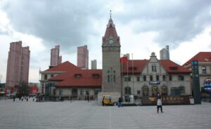 Foto von einem Gebäude mit großem Uhrturm auf einem leeren Platz. Im Hintergrund sind mehrere Hochhäuser zu erkennen.