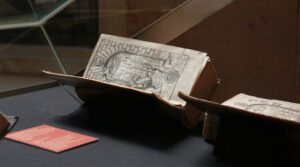 Foto einer geöffneten Vitrine. Darin befinden sich alte, aufgeschlagene Bücher, in denen man Druckgrafiken erkennen kann. neben den Büchern liegt eine kleine rote Tafel.