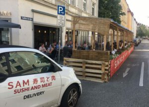 Foto der Außengastronomiefläche in der Gollierstraße. Viele Menschen sitzen vor einem Haus an Tischen zum Essen und Trinken. Vor dem Haus steht ein kleines weißes Auto mit der Aufschrift "SAM Sushi and Meat Delivery".