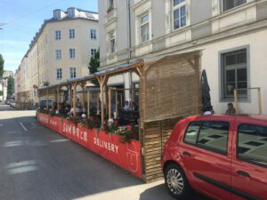 Foto der Außengastronomiefläche einer Straße. Viele Menschen sitzen vor einem Haus an Tischen zum Essen und Trinken. Vor dem Haus steht ein kleines rotes Auto.