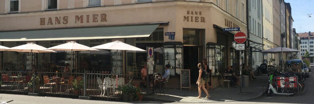 Blick auf einen Häuserblock an einer Straße. An das vorderste Haus wurde "Hans Mier" in Großbuchstaben angebracht. Es handelt sich um eine Art Café mit vielen Schirmen, Stühlen und Tischen draußen.