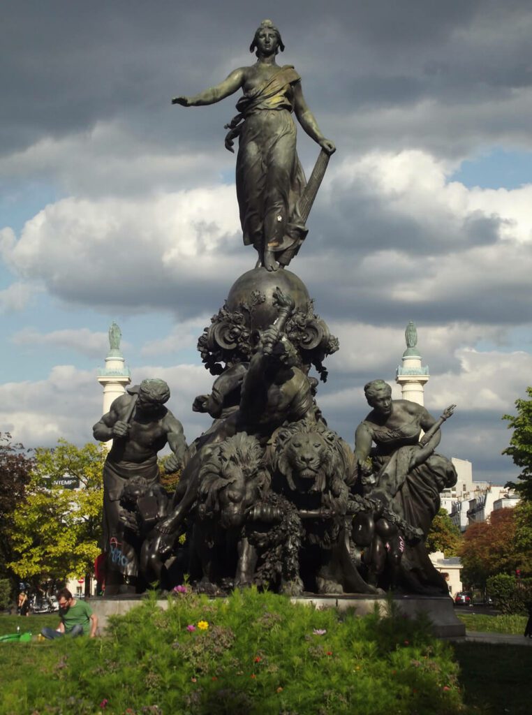 Eine großes Monument aus dunkelgrünem Metall. Die Statue einer Frau mit phrygischer Mütze und Rutenbündel steht auf einem reich dekorierten Wagen der von zwei Löwen gezogen wird. Sie wird von einer weiteren Frau, einem Mann, der eine Fackel in die Höhe reckt und einem muskulösen Mann, der sich einen Hammer auf die Schulter legt, begleitet.