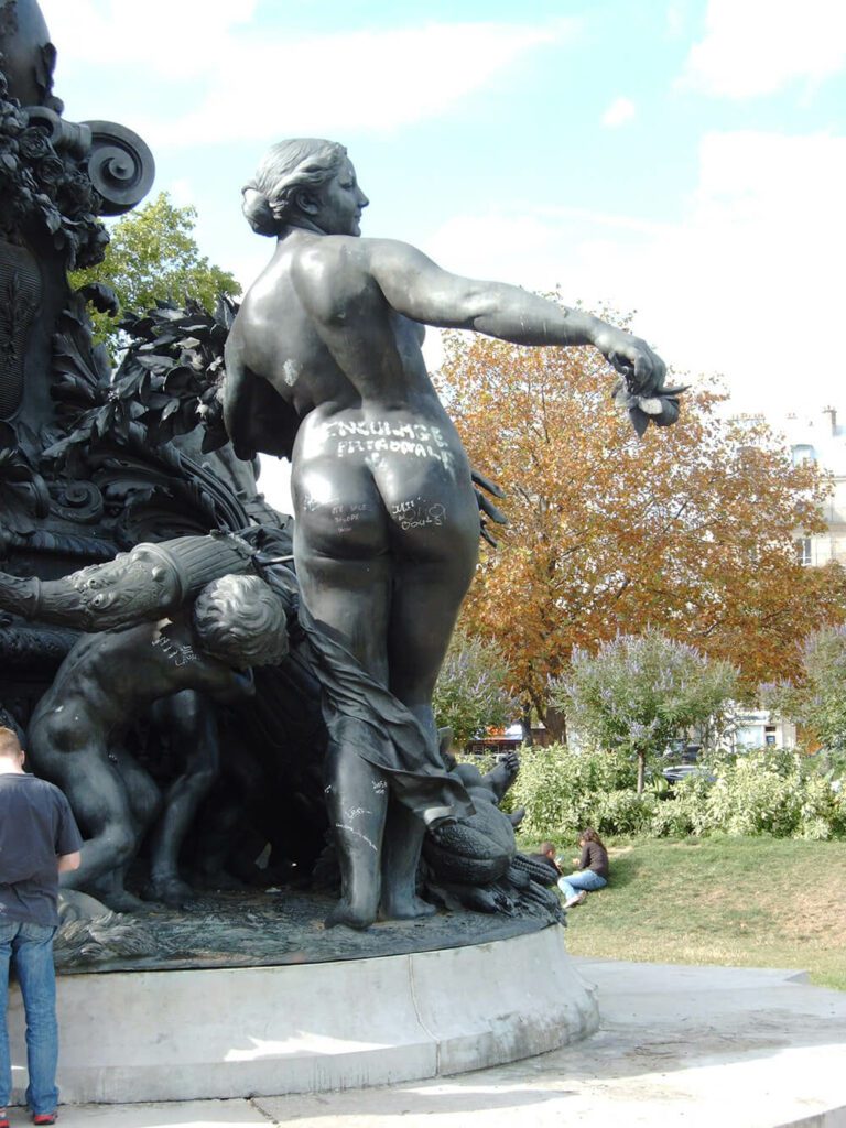 Monumentale Bronzestatue einer nackten Frau mit einer Rose in der Hand. Sie hat uns ihren Rücken zugewandt und ihre beine werden von einer Art Schleier umschlungen. Neben ihr krümmt sich die Statue eines Kindes unter der Last die es auf der Schulter trägt. Beide Statuen wurden mit weißer Farbe beschrieben.