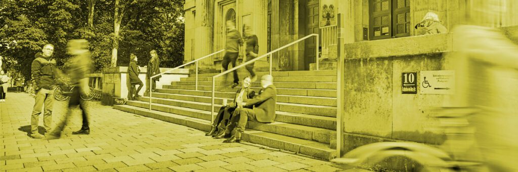 Auf der breiten Treppe vor einem Gebäude gehen, stehen und sitzen Menschen.