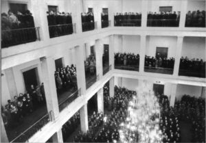 Das Schwarzweißfoto zweigt einen großen, mehrstöckigen Saal, der auf allen Ebenen mit unzähligen Menschen gefüllt ist. In der Mitte des Saales hängt ein imposanter Kronleuchter.