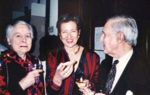 Eine ältere und eine jüngere Frau stehen zusammen mit einem älteren Mann. Alle sind elegant gekleidet und haben Weingläser in der Hand.