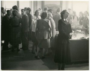 Schwarzweißfoto eines gut gefüllten Raums. Die elegant gekleideten Menschen drängen sich dicht um einen Tisch im Hintergrund. Im Vordergrund steht eine einzelne Frau in schwarzem Blazer und einem langen dunklen Rock.
