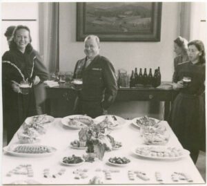 Eine Schwarzweißfotografie eines kleineren, fast glatzköpfigen Mannes in Uniform mit einem Glas in der Hand und einer Frau in einem dunklen Kleid, ebenfalls mit einem Glas in der Hand. Vor ihnen wurde auf einem weißen Tisch ein Buffet hergerichtet, wobei ganz im Vordergrund das Wort "Austria" aus Essen geformt wurde. Im Hintergrund steht ein schwerer Holztisch mit vielen Flaschen und Gläsern.