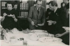 Viele elegant gekleidete Menschen stehen um einen weißen Tisch herum, auf dem ein Essensbuffet eingerichtet wurde. Im Hintergrund steht ein großer Schrank mit vielen Dokumentenordnern.