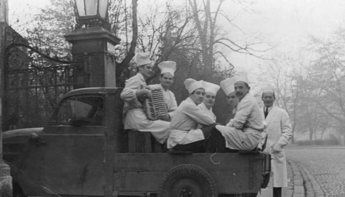 Schwarzweißfoto von Sechs Männer in Kochuniform auf der offenen Ladefläche eines kleinen stehenden Autos neben einer gepflasterten Straße. Ein weiterer Mann ohne Kochmütze steht daneben.