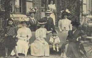 Eine Schwarzweißfotografie von neun Personen, die vor einem Haus stehen. Einige von ihnen sitzen auf einer Bank, andere stehen. Viele tragen auffällige Hüte.