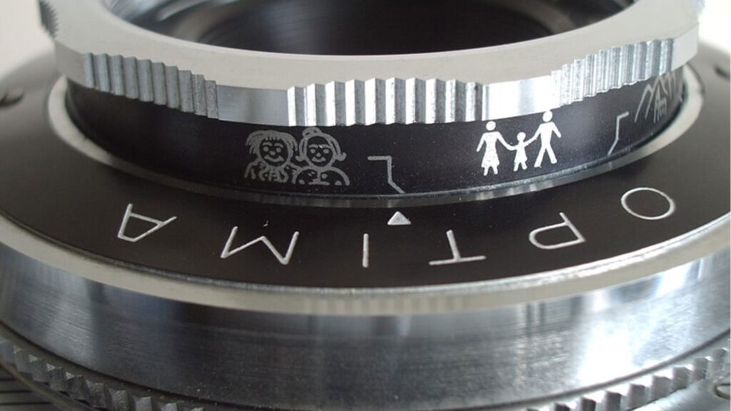 Nahaufnahme eines silbernen Kameraobjektivs mit dem Schriftzug 'OPTIMA' und kleinen weißen Piktogrammen von Familienmitgliedern auf dem Einstellring.