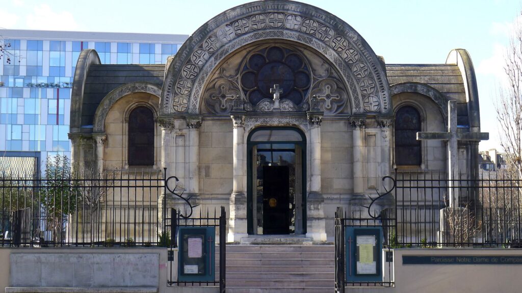 Hinter einem spitzen Eisenzaun, liegt der Eingang zu einer Kirche. Er wird links und rechts von Halbsäulen begrenzt und von einem Kreuz bekrönt. Vorne rechts im Bild, steht ebenfalls ein großes Kreuz aus Stein.