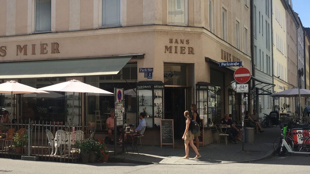 Blick auf einen Häuserblock an einer Straße. An das vorderste Haus wurde "Hans Mier" in Großbuchstaben angebracht. Es handelt sich um eine Art Café mit vielen Schirmen, Stühlen und Tischen draußen.