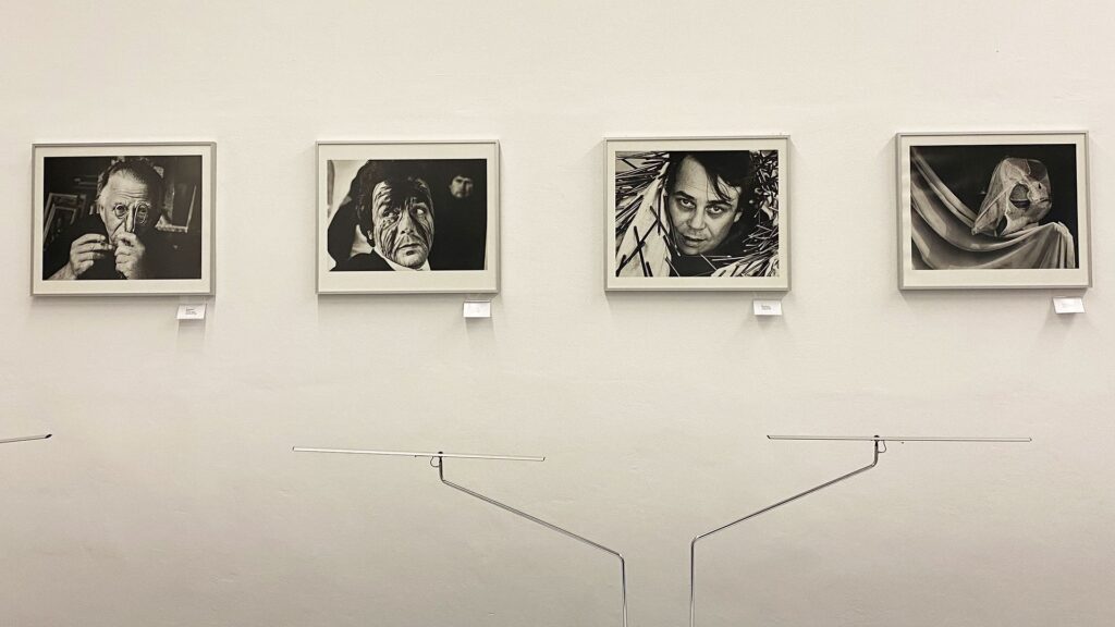 Vier gerahmte Schwarzweißfotografien hängen nebeneinander and einer weißen Wand. Zu sehen sind ein Mann, der sich eine Schere vors Gesicht hält, ein Mann mit verschmiertem Gesicht, ein Mann, der aus einem Tuch mit Nägeln hervorschaut und ein Objekt auf einem hellen Vorhang. Unter den Fotos ist jeweils ein kleiner Zettel angebracht.