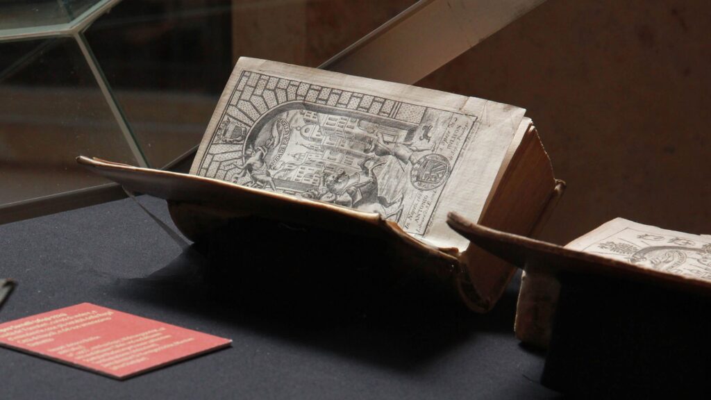 Blick auf einen Umgang um einen Lichthof. Im Gang stehen zahlreiche Vitrinen mit aufgeschlagenen Büchern. An dem Pfeiler direkt in der Bildmitte ist eine große Tafel mit Bild und Text angebracht.