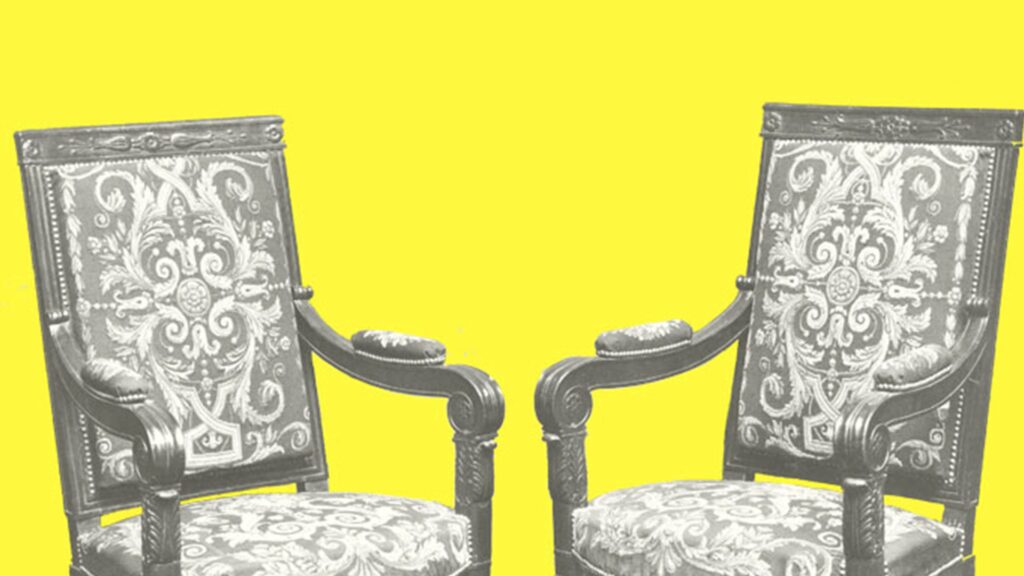 Zwei reich verzierte Holzstühle vor einem zitronengelben Hintergrund. Die Polster der Stühle sind mit aufwendigen Ornamenten bestickt.