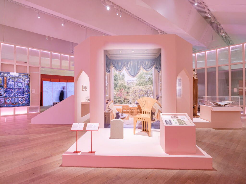 Ausstellungsszene mit einem hellen Holzstuhl vor einem Fenster mit blauen Vorhängen, umgeben von Ausstellungstafeln und Vitrinen in einem modernen Museum.