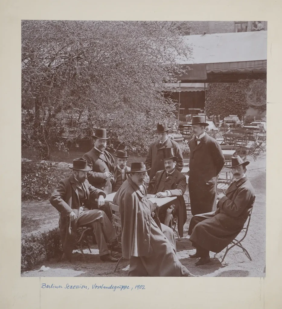 Gruppe von acht Männern in historischer Kleidung sitzt und steht um einen Gartentisch in einem Außenbereich mit Bäumen und Stühlen im Hintergrund