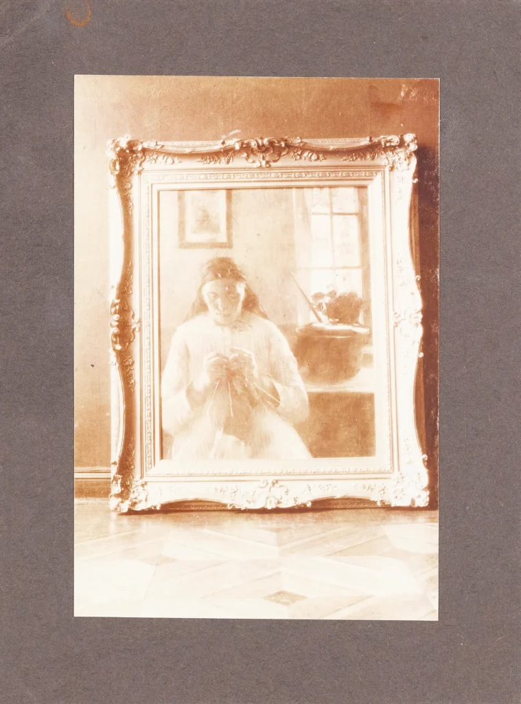 Porträt einer sitzenden Person in einem weißen Kleid, die in einem großen, verzierten Spiegel vor einem Fenster mit Pflanzen reflektiert wird