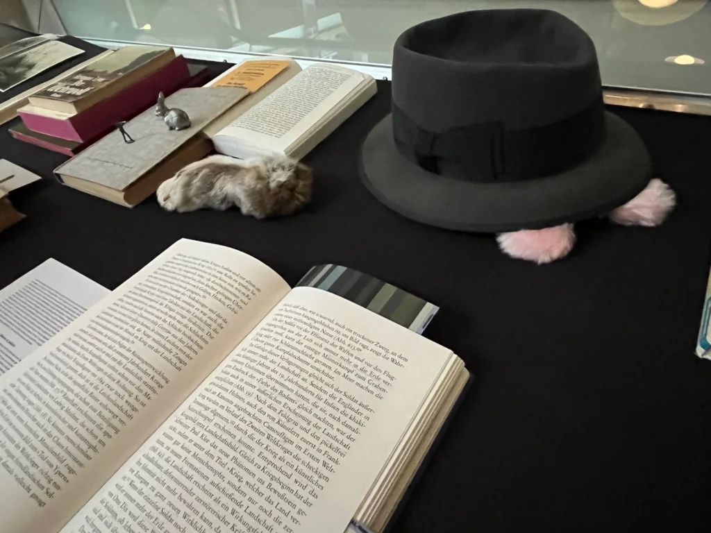 Auf schwarzem Tuch liegende Bücher, ein grauer Hut unter welchem rosa Plüschohren hervorschauen und ein Fellstück