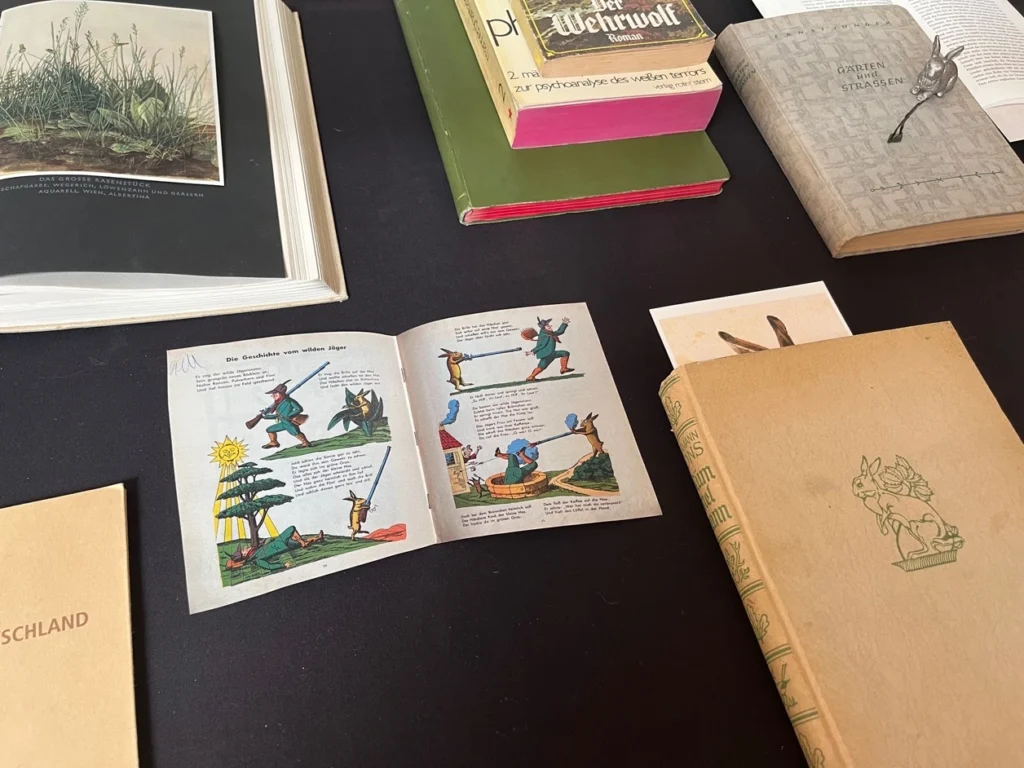 Mehrere aufgeschlagene und geschlossene Bücher liegen auf einem schwarzen Tuch darunter eine aufgeschlagene illustrierte Doppelseite mit einer Geschichte über einen Jäger und Tiere