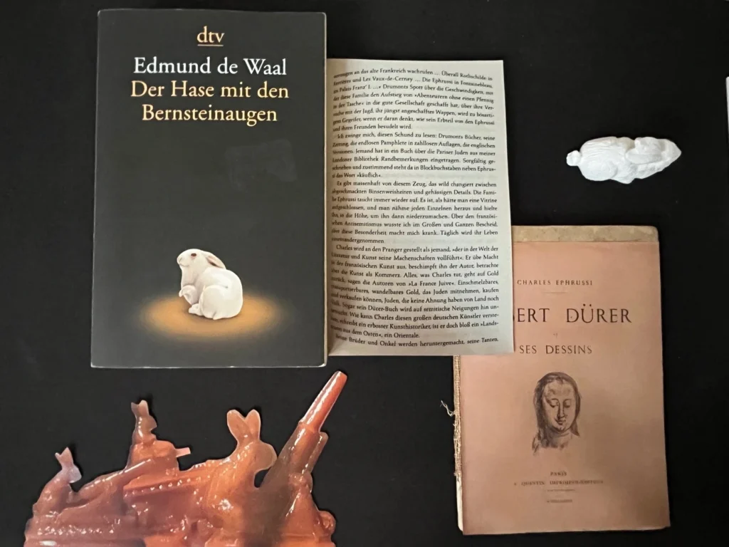 Buch "Der Hase mit den Bernsteinaugen" von Edmund de Waal, ein aufgeschlagenes Buch mit Text, ein Buch über Albrecht Dürer von Charles Ephrussi und mehrere kleine Hasenfiguren aus verschiedenen Materialien auf schwarzem Hintergrund