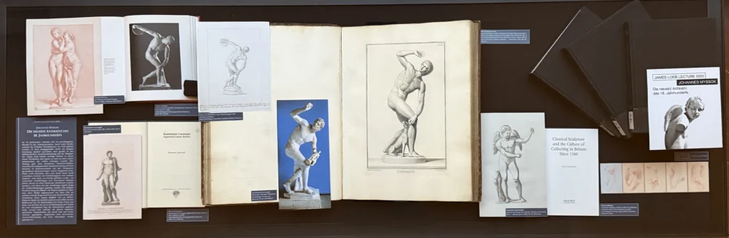 Graue Statue eines nackten Mannes in einer dynamischen Pose mit einer Scheibe in der Hand vor gelbem Hintergrund