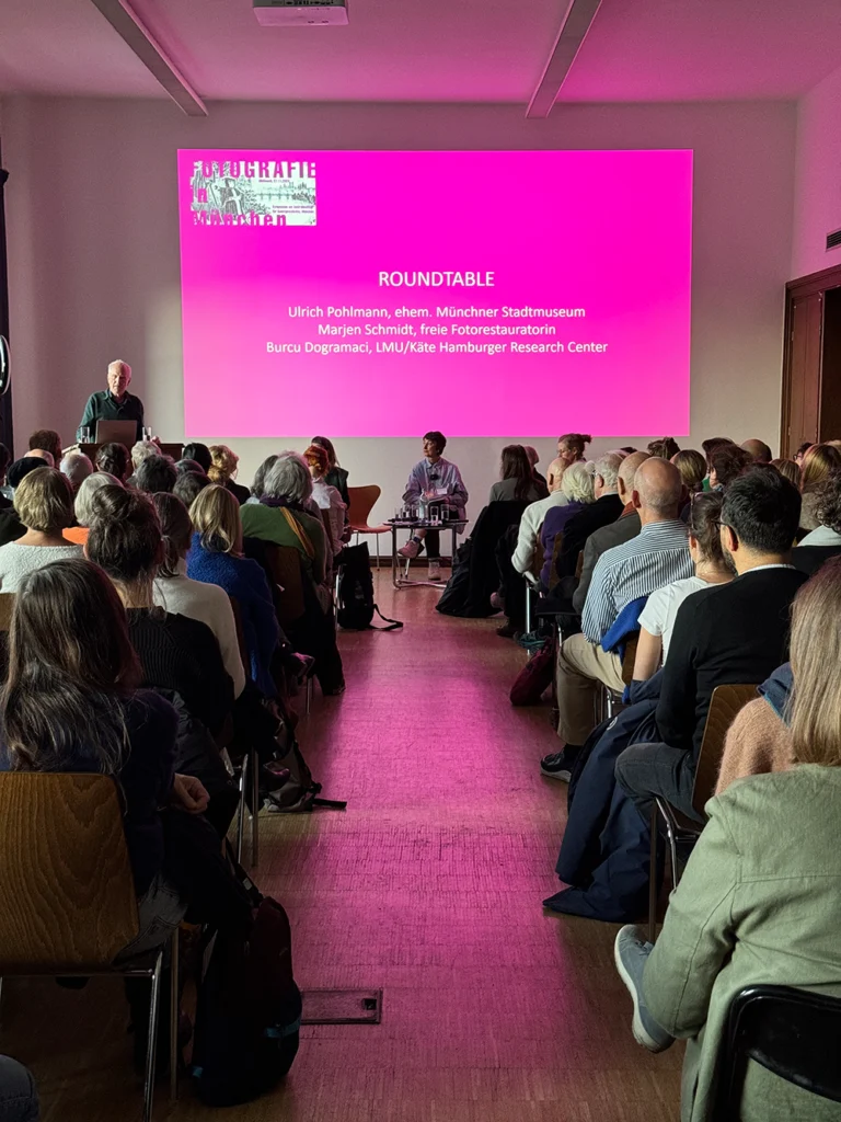 Publikum sitzt in einem Raum vor einer pink beleuchteten Leinwand mit dem Titel 'ROUNDTABLE' und mehreren Namen darunter