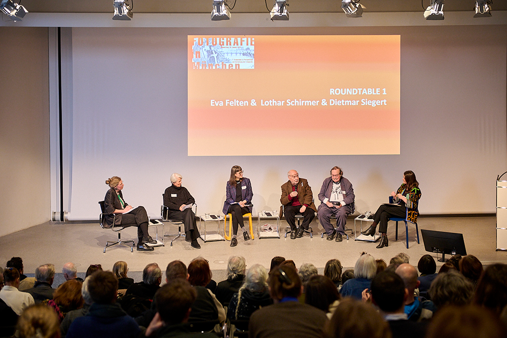 Sechs Personen sitzen auf Stühlen auf einer Bühne vor einem Publikum, im Hintergrund eine Leinwand mit dem Text 'ROUND TABLE 1 Eva Felten & Lothar Schirmer & Dietmar Siegert'